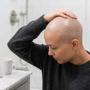Une personne au crâne rasé se touche la tête dans une salle de bain avec des produits de rasage sur le comptoir.