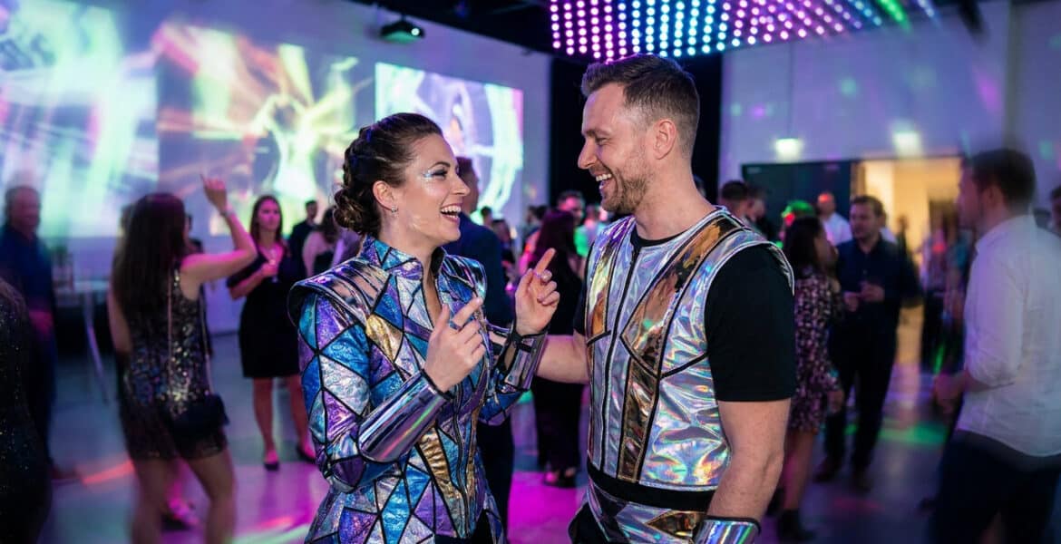 Un couple riant lors d'une fête, portant des costumes futuristes brillants et métalliques.