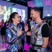 Un couple riant lors d'une fête, portant des costumes futuristes brillants et métalliques.