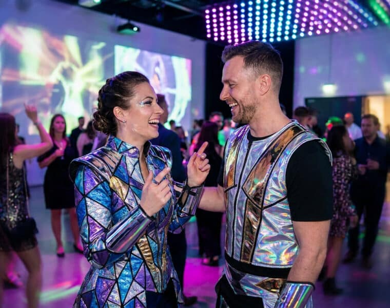 Un couple riant lors d'une fête, portant des costumes futuristes brillants et métalliques.