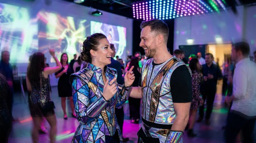Un couple riant lors d'une fête, portant des costumes futuristes brillants et métalliques.