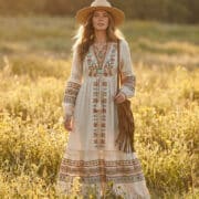 Une femme blonde portant une robe longue bohème, un chapeau de paille et un sac à franges dans un champ de fleurs.