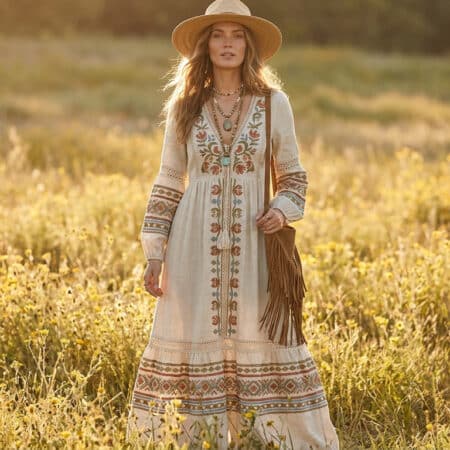 Une femme blonde portant une robe longue bohème, un chapeau de paille et un sac à franges dans un champ de fleurs.