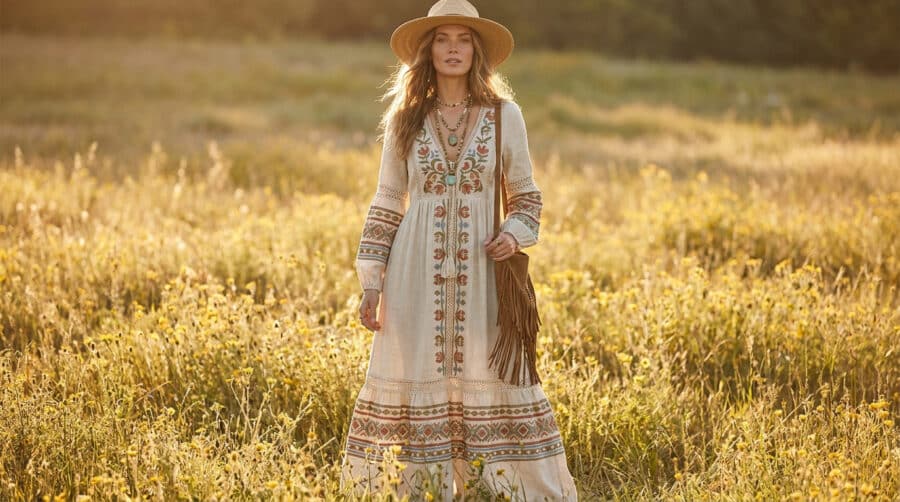 Une femme blonde portant une robe longue bohème, un chapeau de paille et un sac à franges dans un champ de fleurs.