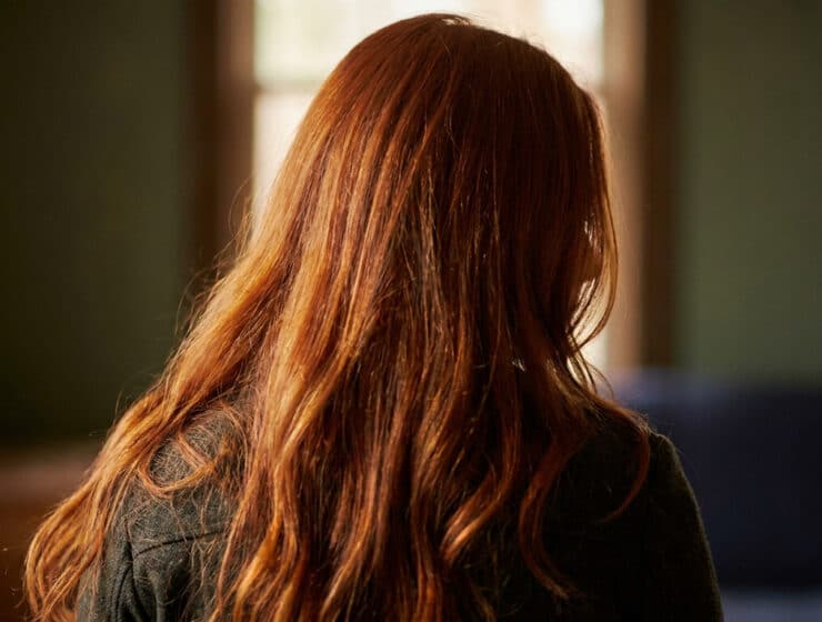 Vue de dos d'une femme aux longs cheveux roux brillants éclairés par la lumière naturelle d'une fenêtre.
