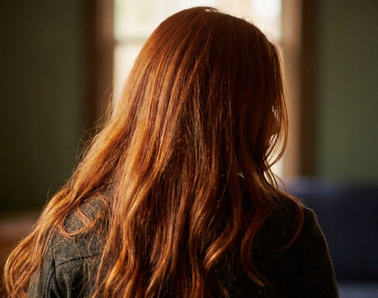 Vue de dos d'une femme aux longs cheveux roux brillants éclairés par la lumière naturelle d'une fenêtre.