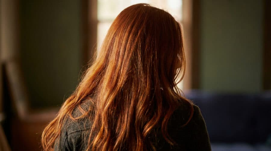 Vue de dos d'une femme aux longs cheveux roux brillants éclairés par la lumière naturelle d'une fenêtre.