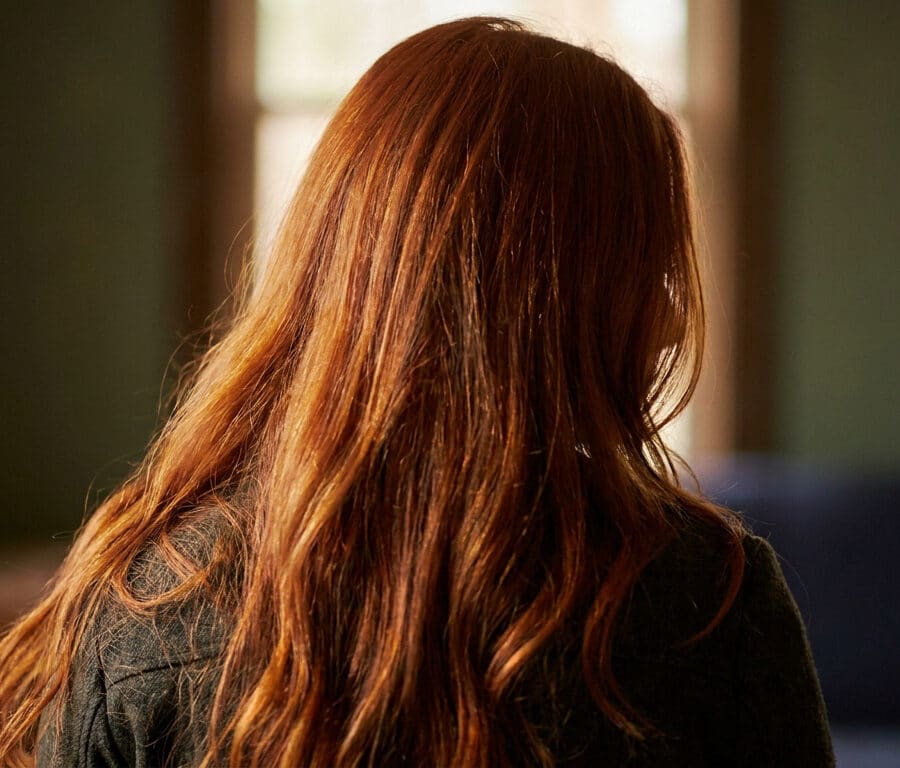 Vue de dos d'une femme aux longs cheveux roux brillants éclairés par la lumière naturelle d'une fenêtre.