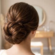 Vue arrière d'une femme avec un chignon brun élégant, dans une chambre lumineuse avec une coiffeuse en arrière-plan.