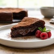 Une part de gâteau au chocolat fondant saupoudré de cacao, décorée de framboises et de menthe sur une assiette blanche.