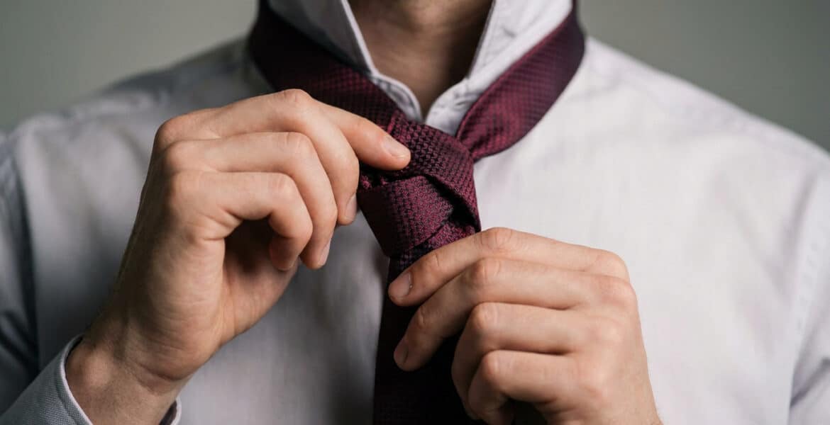 Gros plan sur les mains d'un homme ajustant une cravate bordeaux texturée sur une chemise blanche.