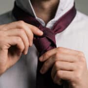 Gros plan sur les mains d'un homme ajustant une cravate bordeaux texturée sur une chemise blanche.