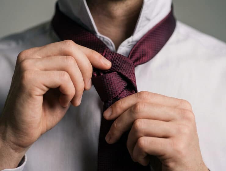 Gros plan sur les mains d'un homme ajustant une cravate bordeaux texturée sur une chemise blanche.