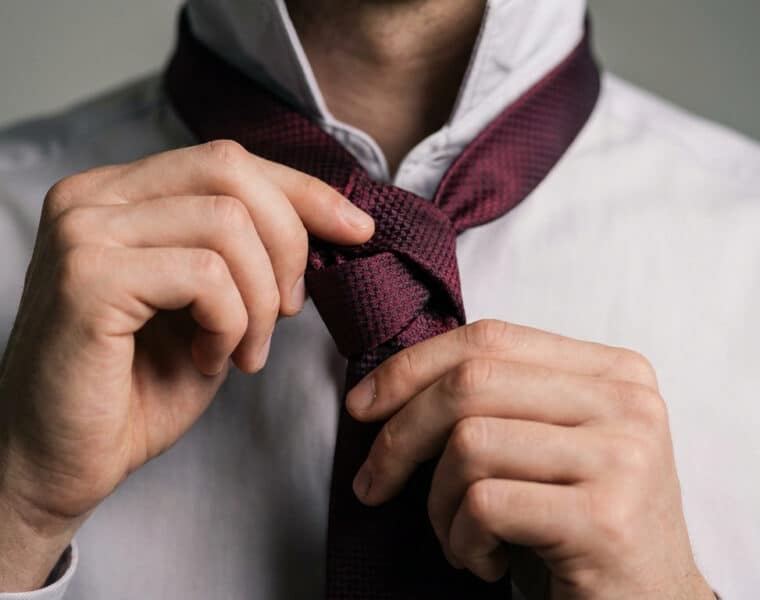 Gros plan sur les mains d'un homme ajustant une cravate bordeaux texturée sur une chemise blanche.