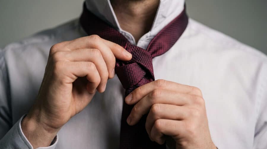 Gros plan sur les mains d'un homme ajustant une cravate bordeaux texturée sur une chemise blanche.