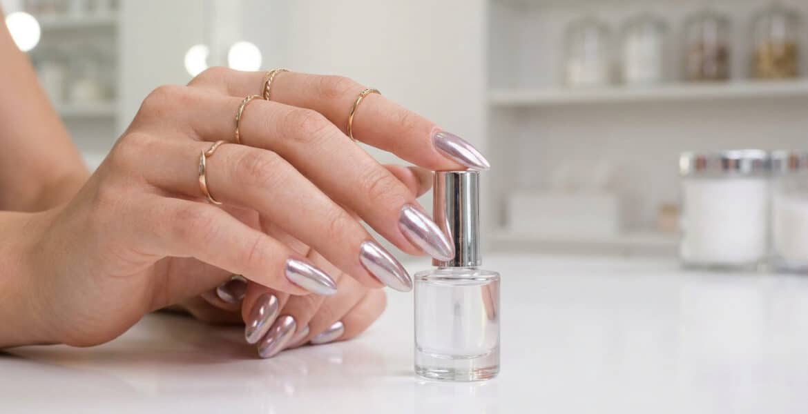 Main féminine avec des ongles longs en amande et un fini effet miroir chromé, tenant un flacon de vernis transparent.