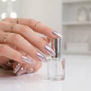 Main féminine avec des ongles longs en amande et un fini effet miroir chromé, tenant un flacon de vernis transparent.