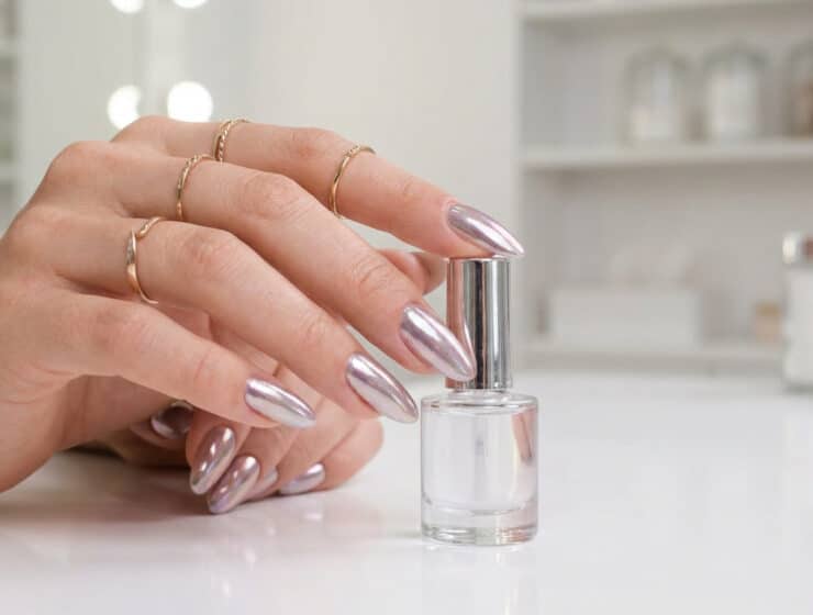 Main féminine avec des ongles longs en amande et un fini effet miroir chromé, tenant un flacon de vernis transparent.