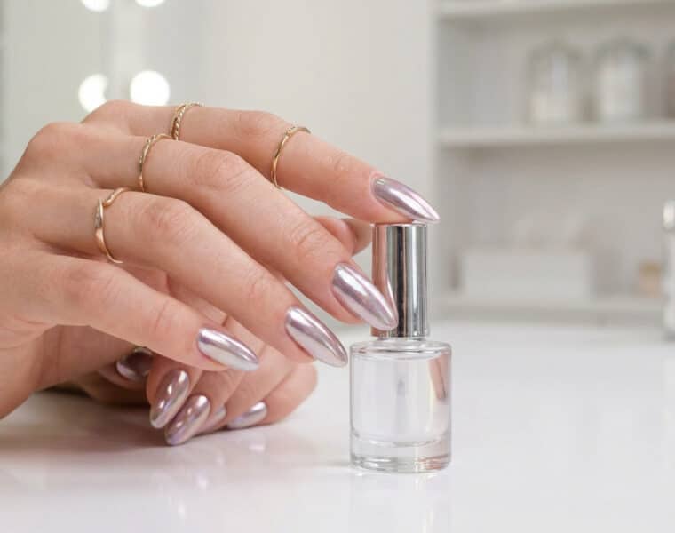Main féminine avec des ongles longs en amande et un fini effet miroir chromé, tenant un flacon de vernis transparent.