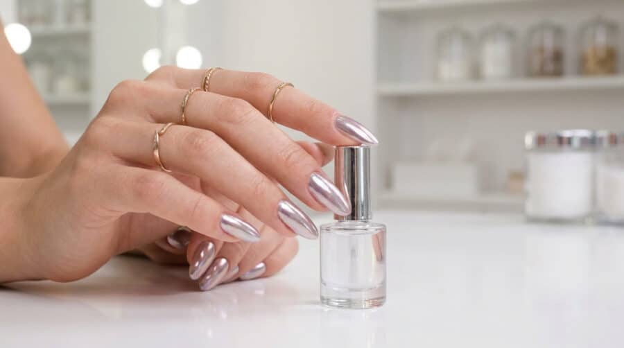 Main féminine avec des ongles longs en amande et un fini effet miroir chromé, tenant un flacon de vernis transparent.