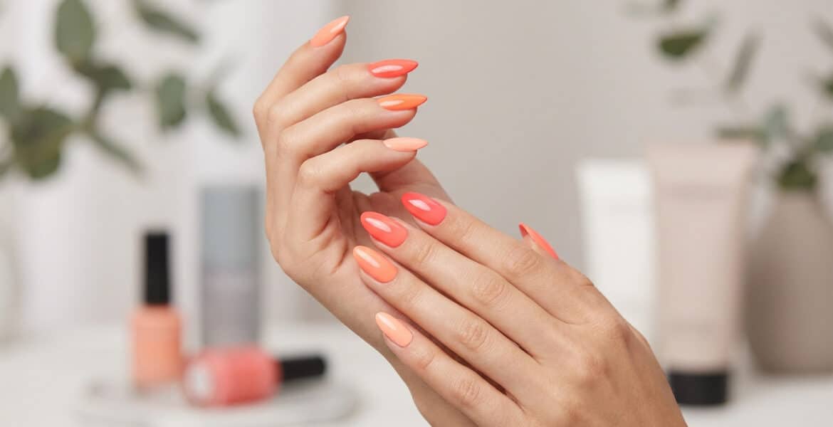 Mains avec une manucure aux différentes nuances de corail, avec des flacons de vernis en arrière-plan flou.