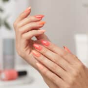 Mains avec une manucure aux différentes nuances de corail, avec des flacons de vernis en arrière-plan flou.