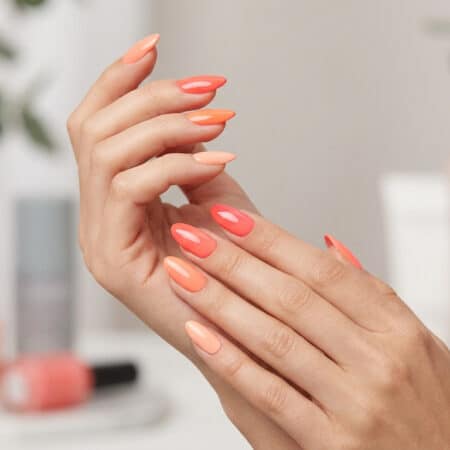 Mains avec une manucure aux différentes nuances de corail, avec des flacons de vernis en arrière-plan flou.
