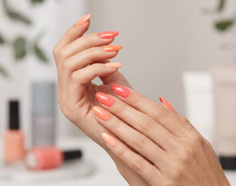 Mains avec une manucure aux différentes nuances de corail, avec des flacons de vernis en arrière-plan flou.