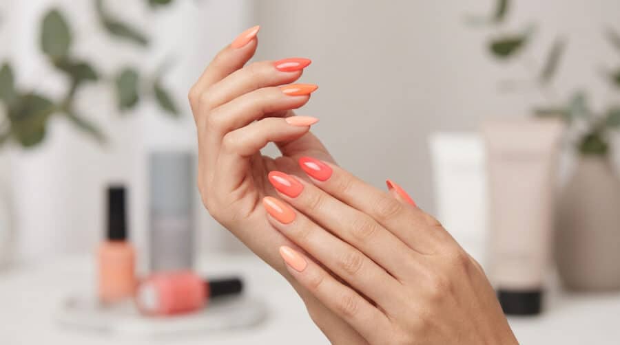Mains avec une manucure aux différentes nuances de corail, avec des flacons de vernis en arrière-plan flou.