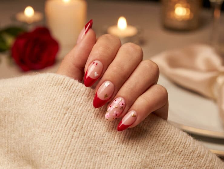 Une main portant un pull beige avec des ongles en amande décorés de motifs rouges, cœurs et fleurs pour la Saint-Valentin.