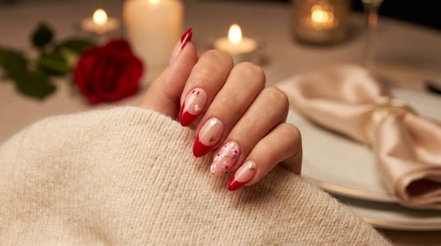 Une main portant un pull beige avec des ongles en amande décorés de motifs rouges, cœurs et fleurs pour la Saint-Valentin.
