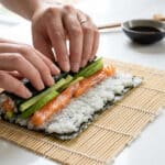 Mains en gros plan roulant un maki avec du saumon, de l'avocat et du riz sur une natte en bambou.
