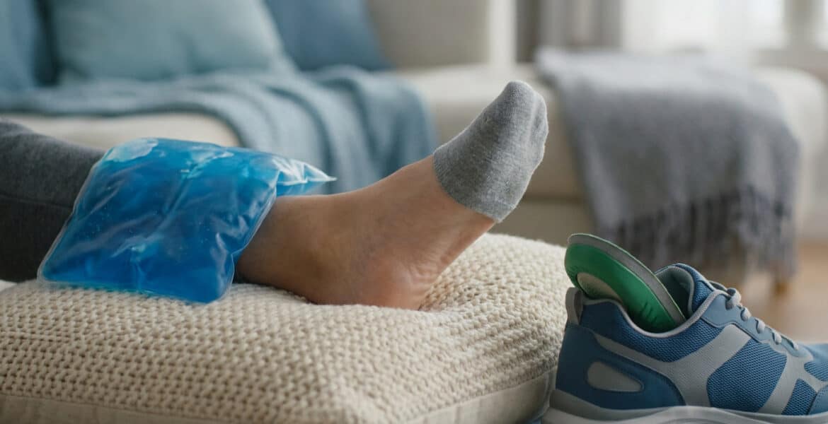 Un pied reposant sur un coussin avec une poche de glace bleue, une chaussure de sport avec semelle orthopédique à côté.