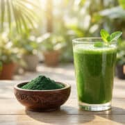 Un verre de smoothie vert et un bol de poudre de spiruline sur une table en bois avec des plantes en arrière-plan.