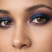 Gros plan sur un regard aux yeux marron sublimé par un maquillage smokey eye dans les tons bleu marine et prune.