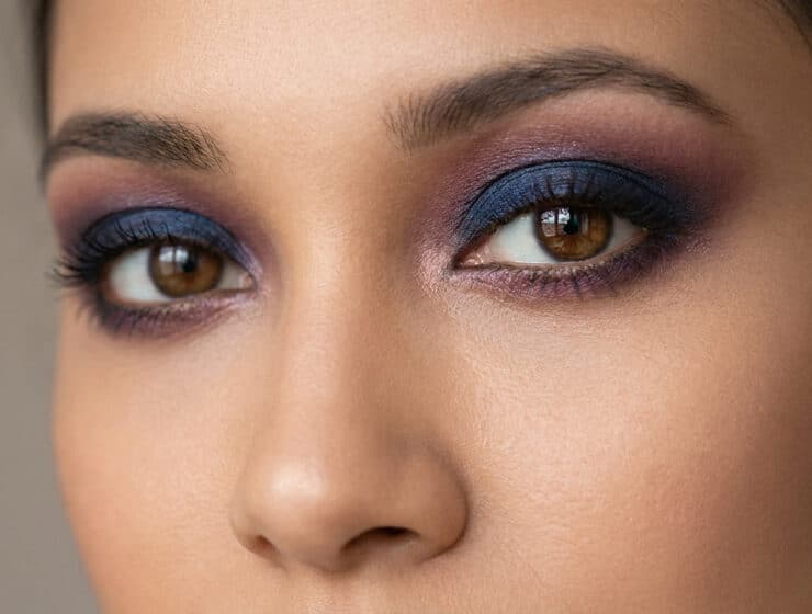 Gros plan sur un regard aux yeux marron sublimé par un maquillage smokey eye dans les tons bleu marine et prune.