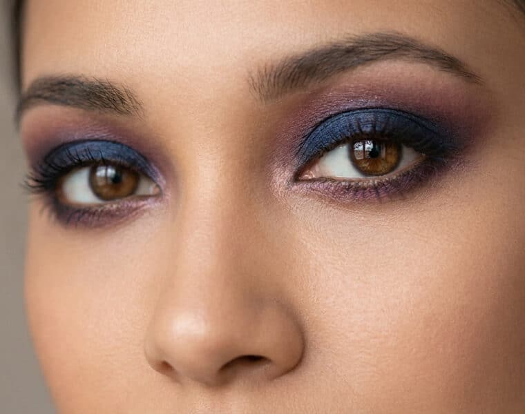 Gros plan sur un regard aux yeux marron sublimé par un maquillage smokey eye dans les tons bleu marine et prune.