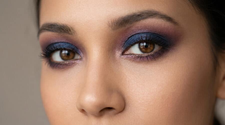 Gros plan sur un regard aux yeux marron sublimé par un maquillage smokey eye dans les tons bleu marine et prune.